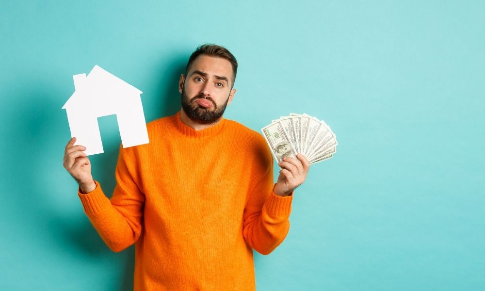 Uomo con in mano dei soldi e una sagoma di casa a riprendere il costo della casa come sinonimo di povertà